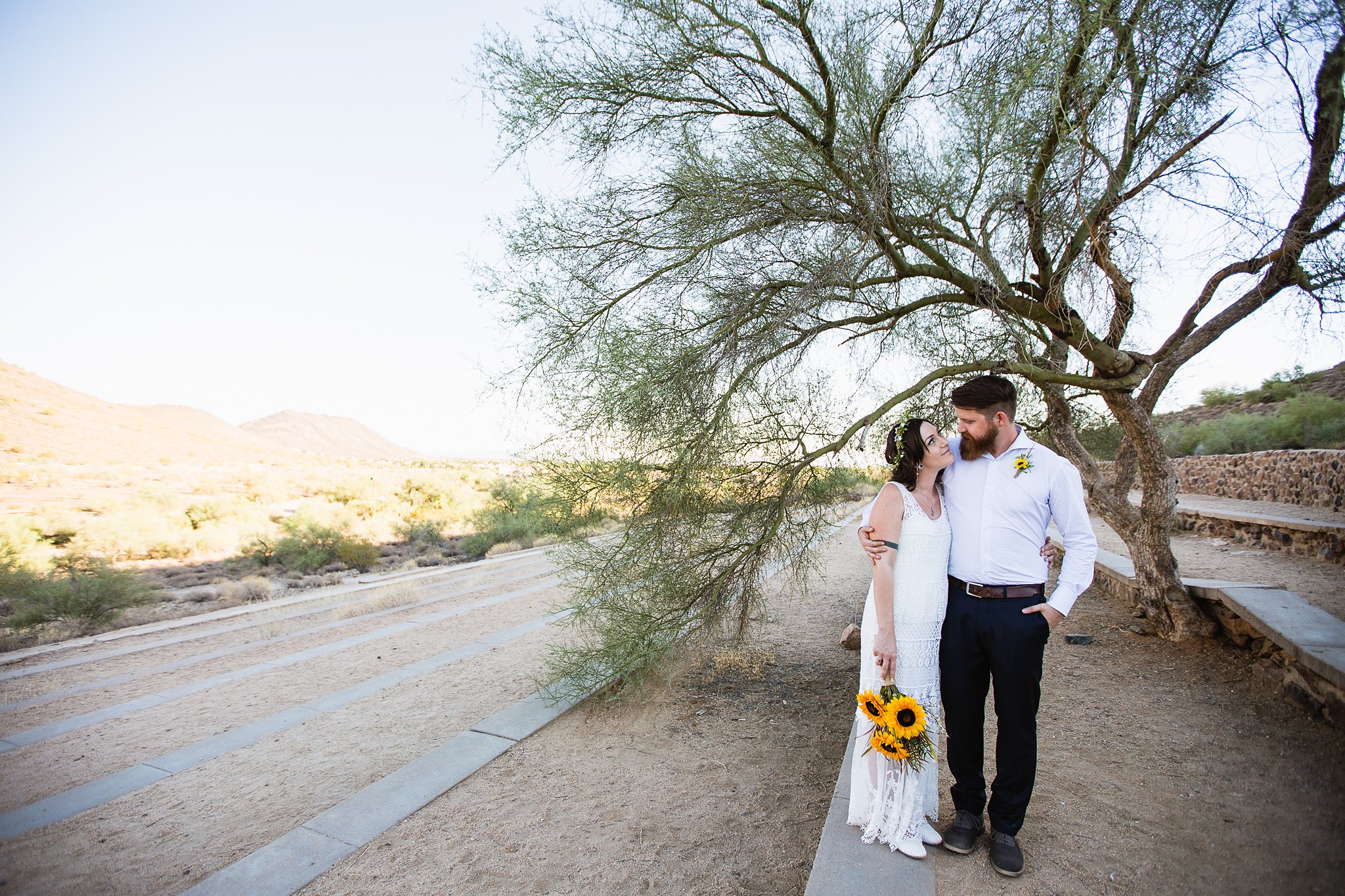 Simple boho inspired bride and groom look at each other in front of a desert tree by Phoenix wedding photographer PMA Photography.