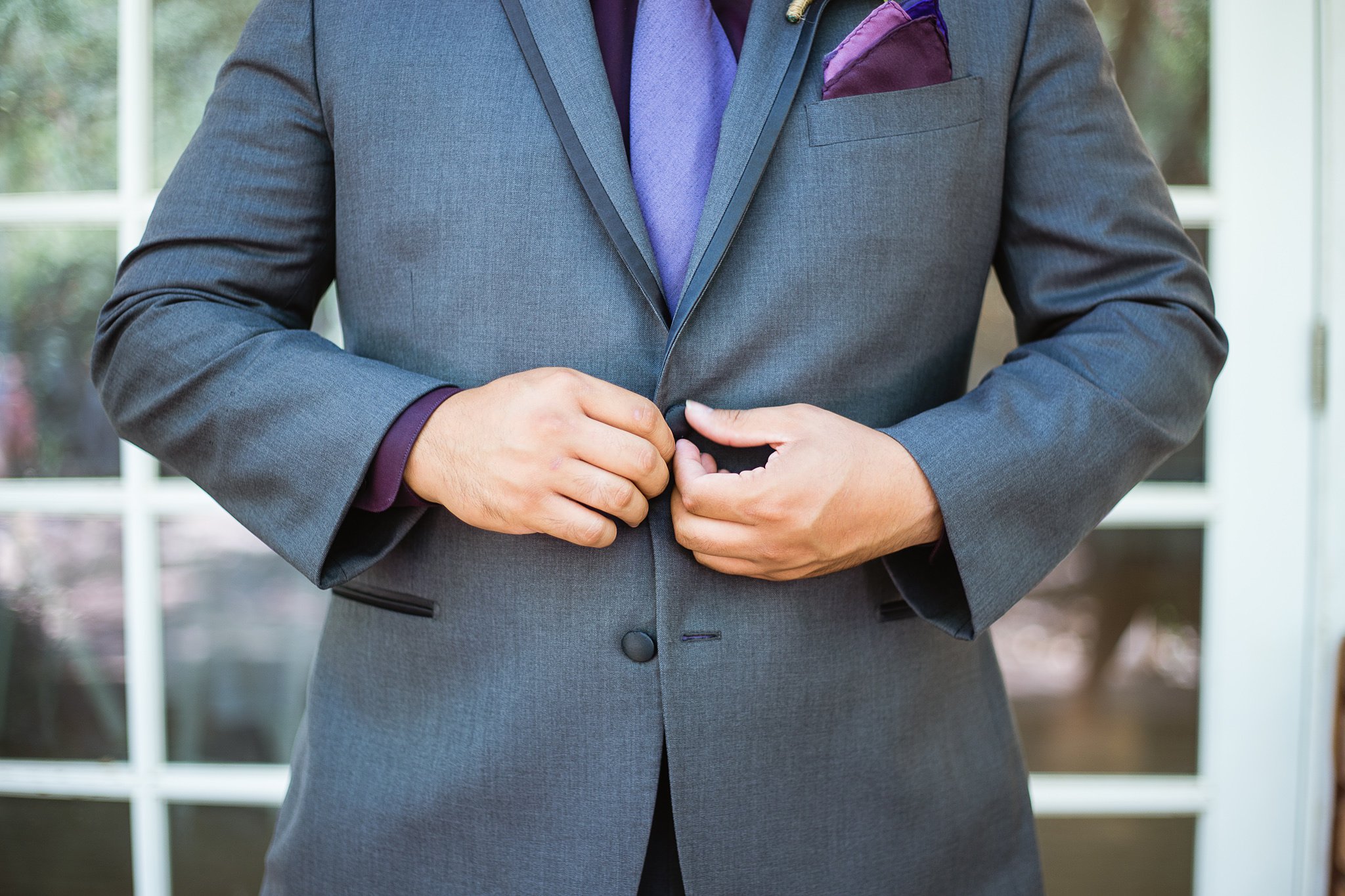 Groom buttoning the jacket of his purple and grey suit while getting ready for his wedding day by Phoenix wedding photographer PMA Photography.