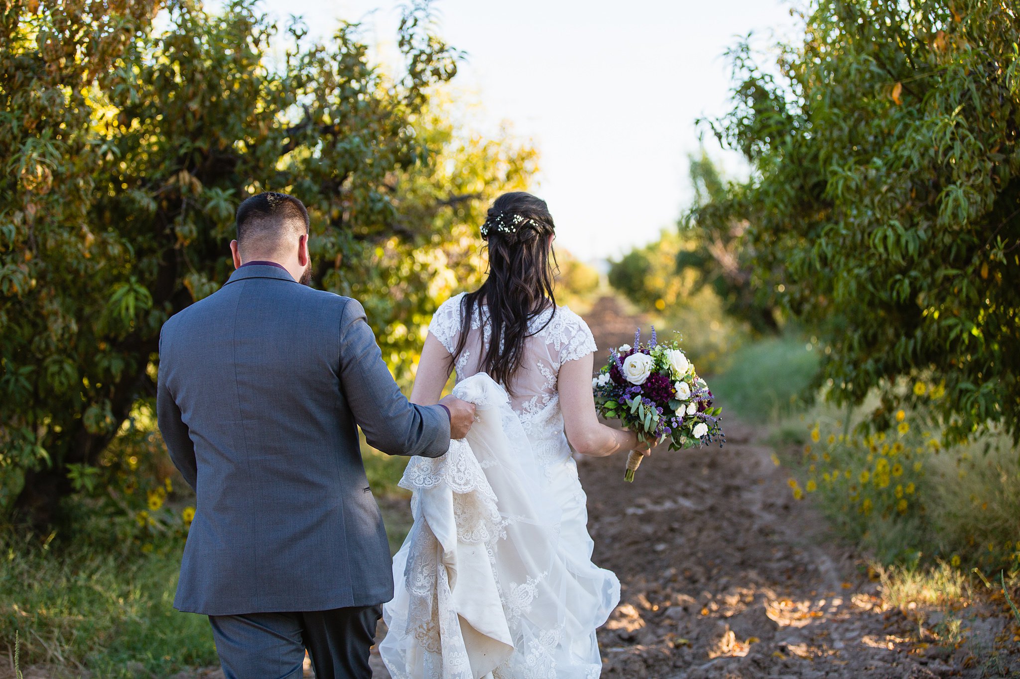Groom helping bride walk through the orchard at the Farmhouse at Schnepf Farms by Phoenix wedding photographer PMA Photography.