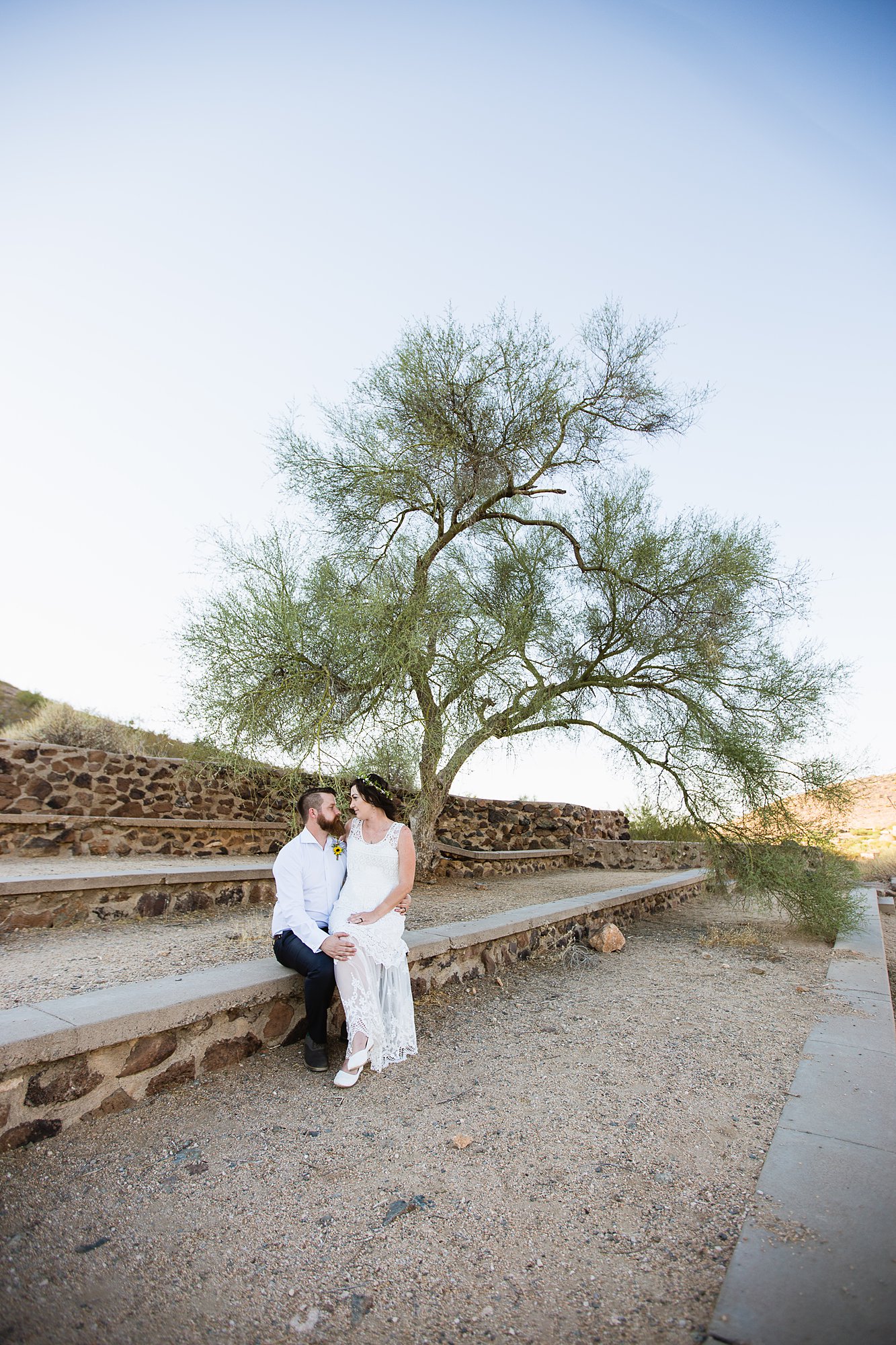 Simple boho bride and groom sitting in front of a tree in the Arizona desert by Phoenix wedding photographers PMA Photography.