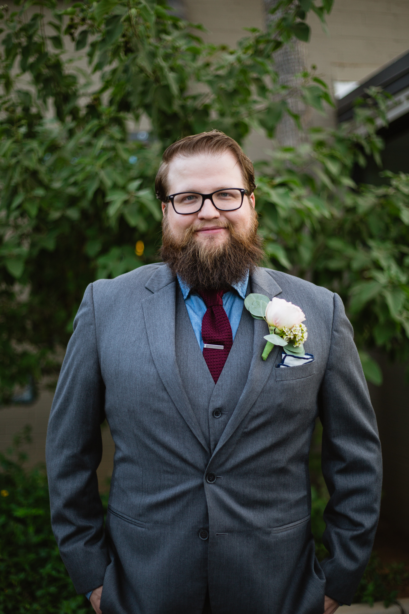 Portrait of groom in his outfit of a grey suit with blue and maroon hints in front of green plants on his wedding day.