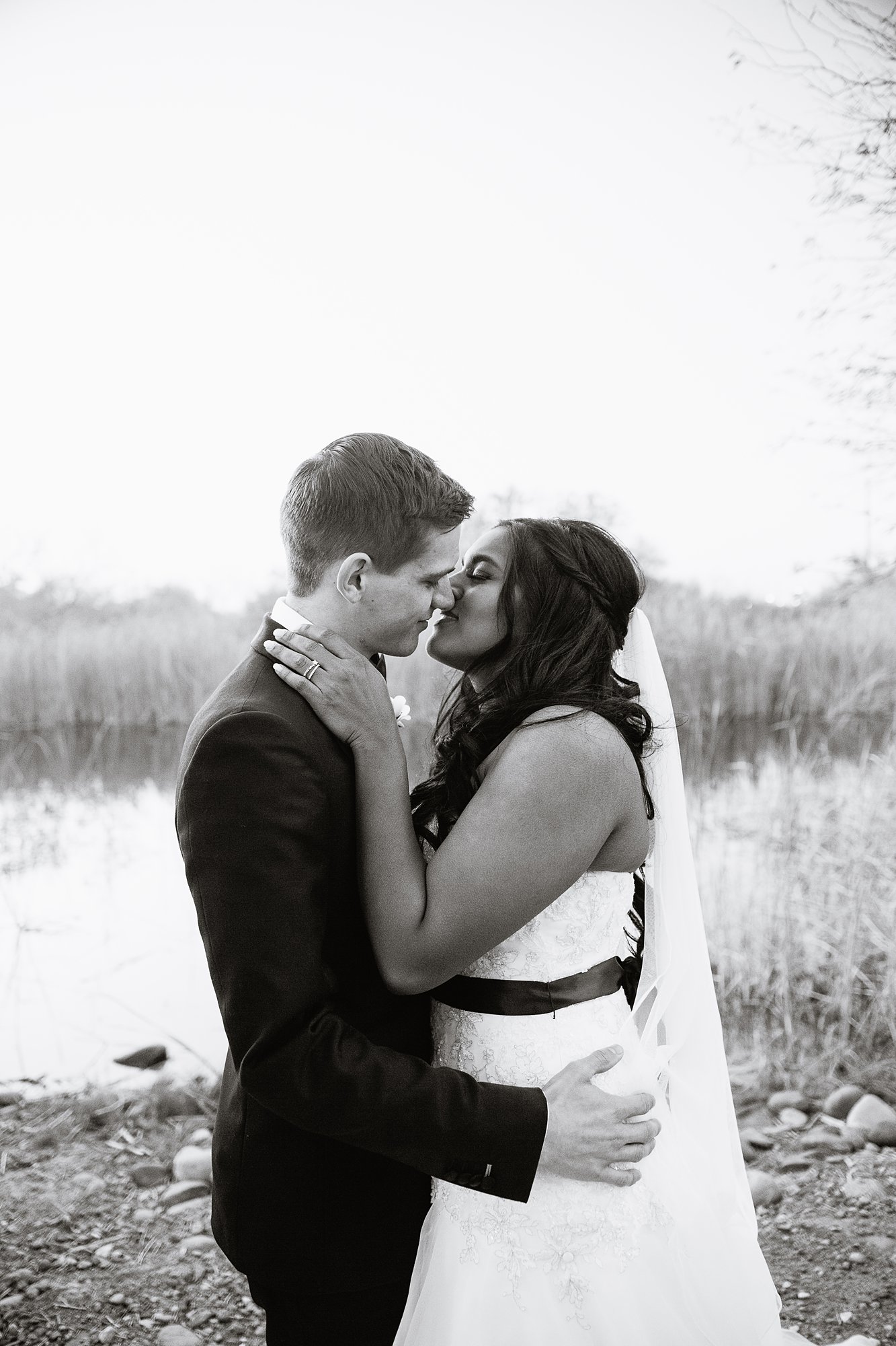 Black and white image of bride and groom sharing a kiss in front of a small lake at the Rio Salado Audubon center by Phoenix wedding photographer PMA Photography.