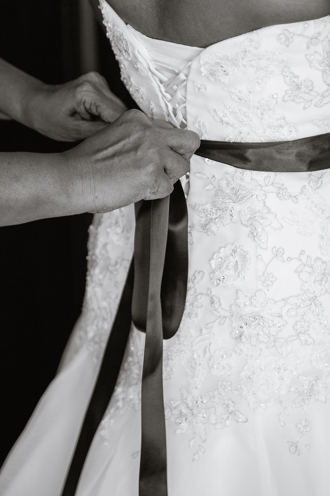 Black and white image of the bride's mom tying a sash on her wedding dress.