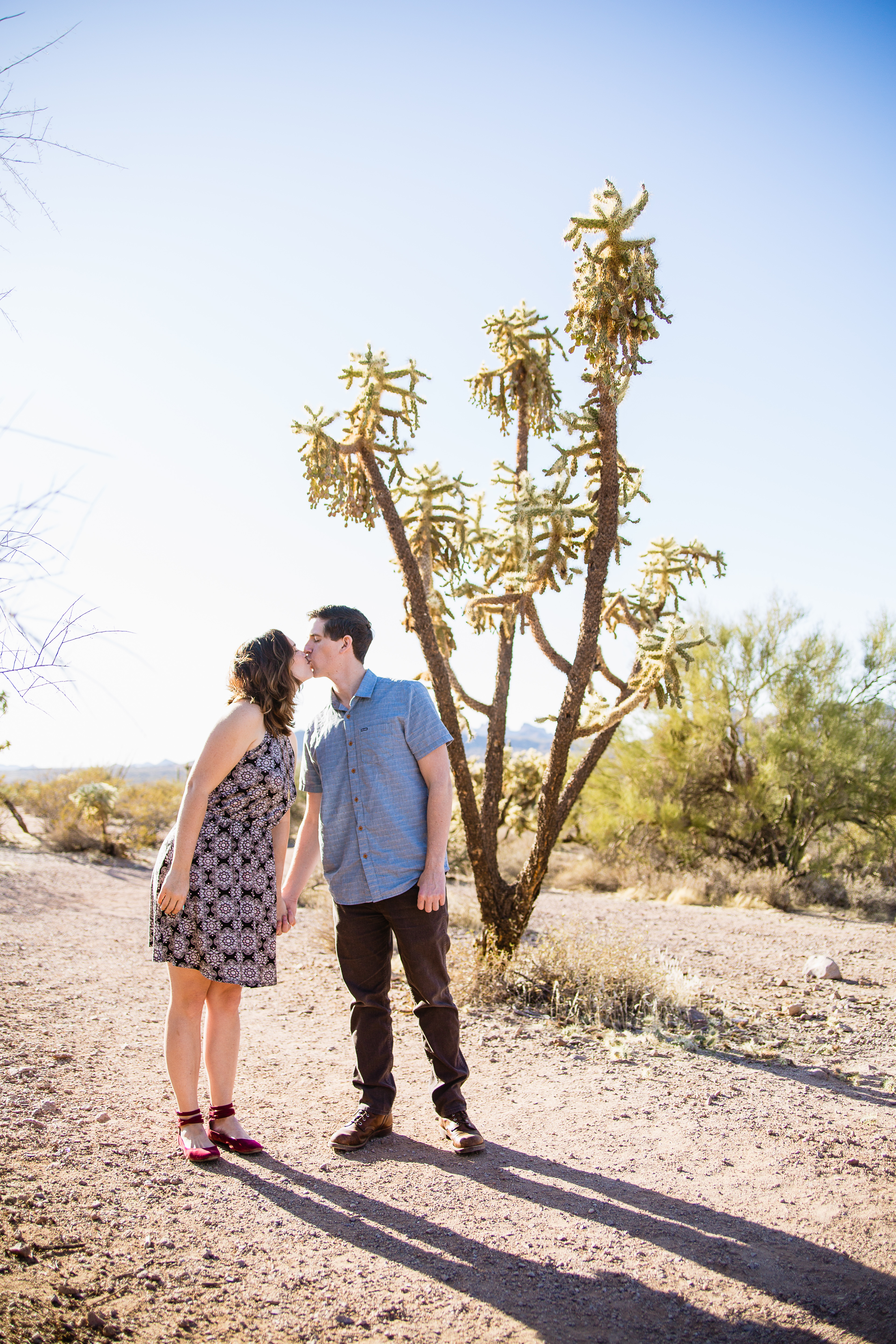 Couple kissing in front of jumping cactus during desert engagement session at Lost Dutchman State Park by Phoenix wedding photographers PMA Photography.