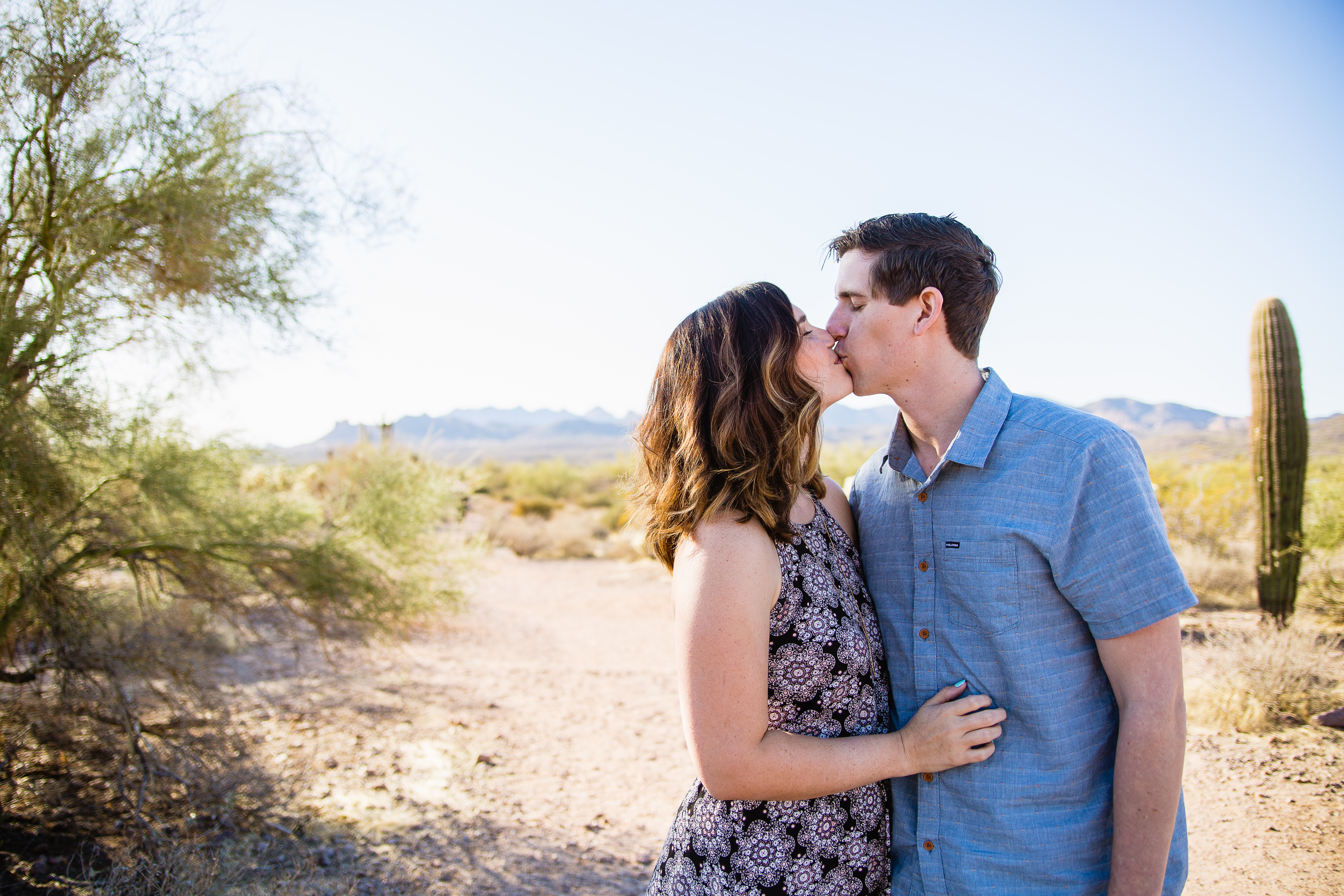 Couple kissing in front of desert mountain backdrop at their engagement session at Lost Dutchman State Park by Phoenix wedding photographers PMA Photography..