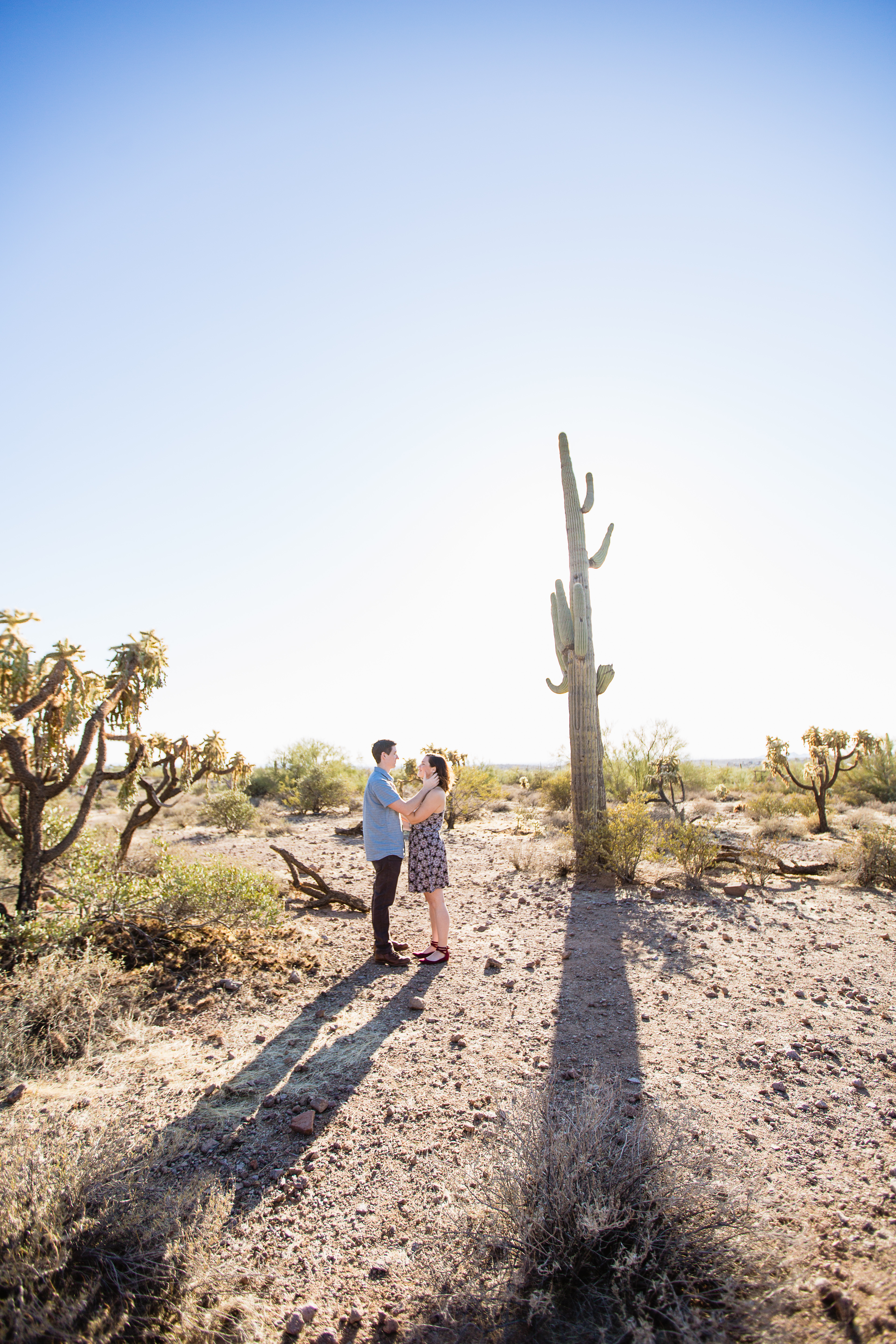 Couple looking at each other in front of large saguaro cactus at their engagement session at Lost Dutchman State Park by Phoenix wedding photographers PMA Photography.