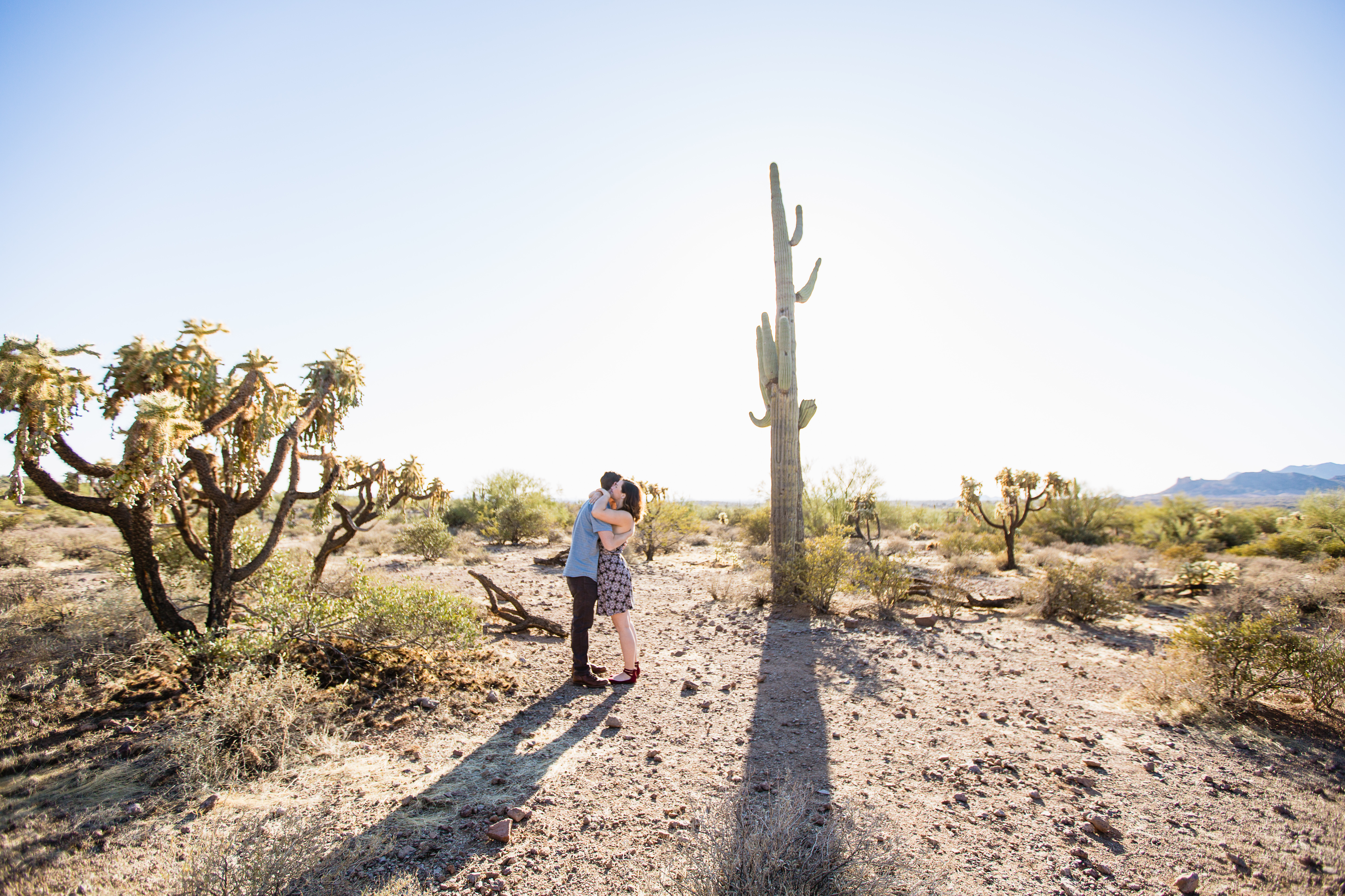 Couple hugging at each other in front of large saguaro cactus at their engagement session at Lost Dutchman State Park by Phoenix wedding photographers PMA Photography.