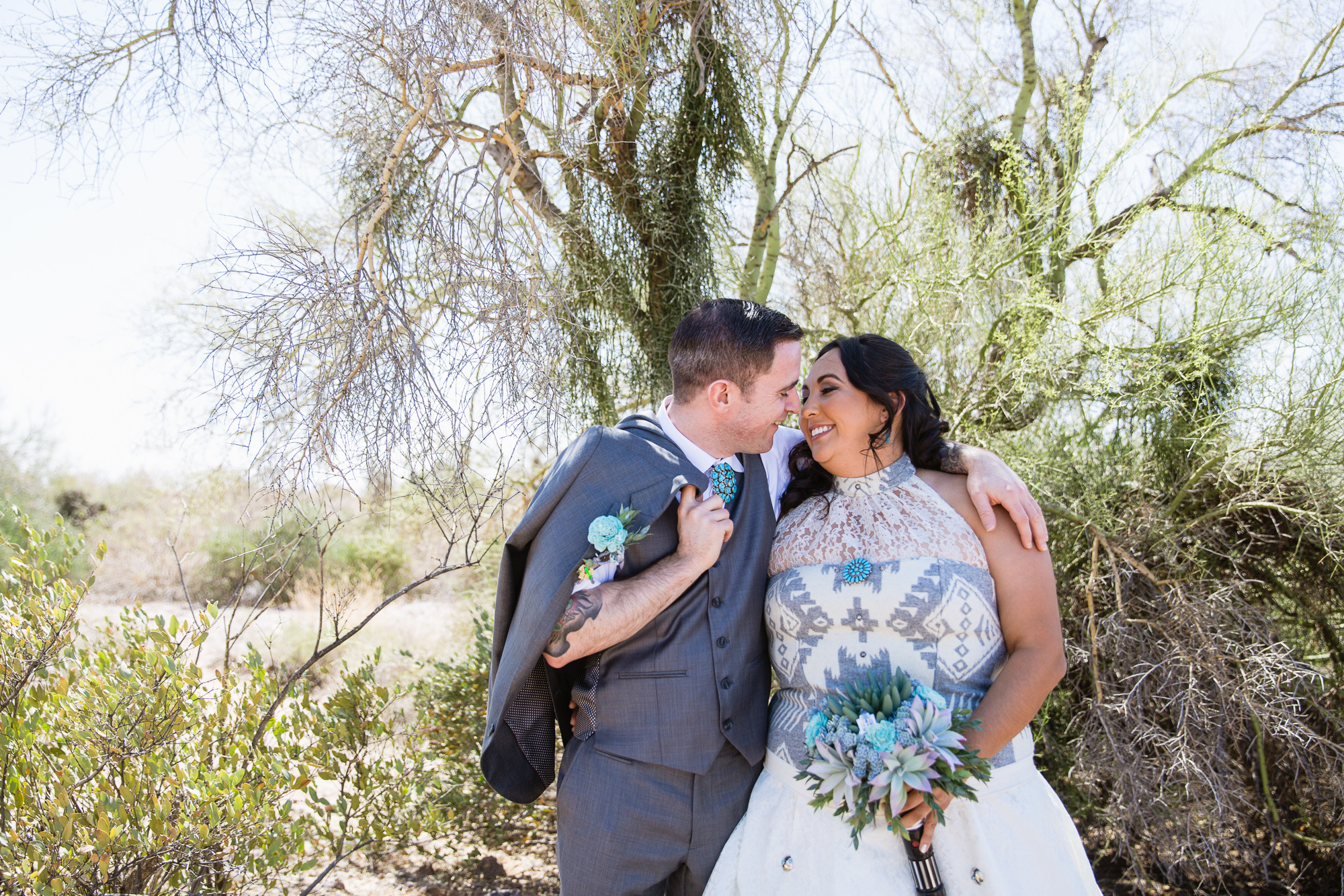 Tattooed and Irish groom and Navajo bride in a native inspired wedding dress share a laugh at their Lost Dutchman State Park Turquoise Star Wars wedding by Phoenix wedding photographers PMA Photography.