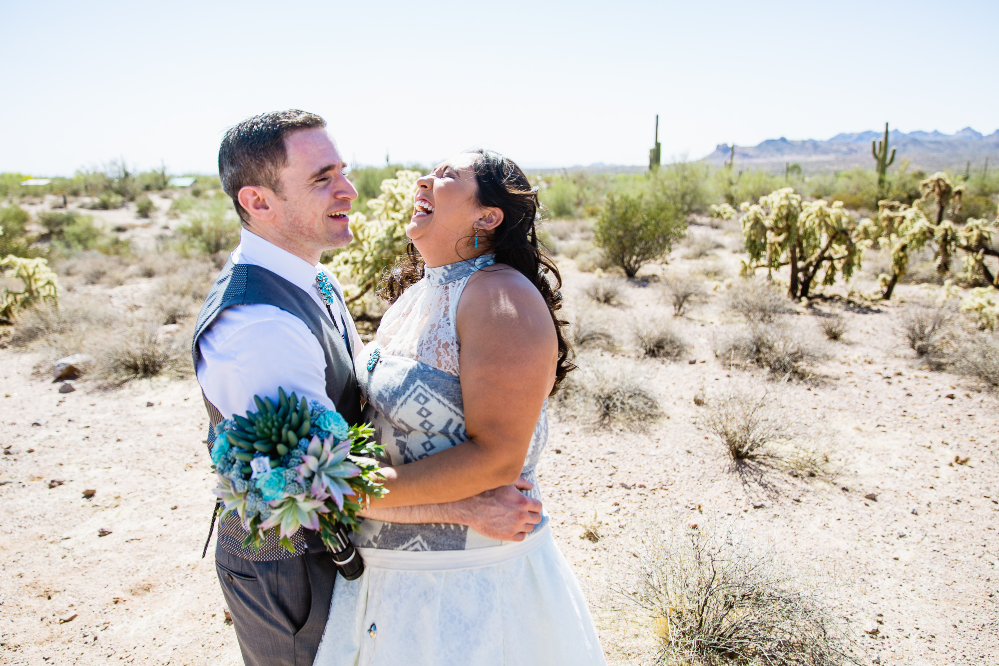 Bride and groom sharing a laugh in turquoise and grey wedding attire in the Arizona desert at Lost Dutchman State Park by wedding photographers PMA Photography.