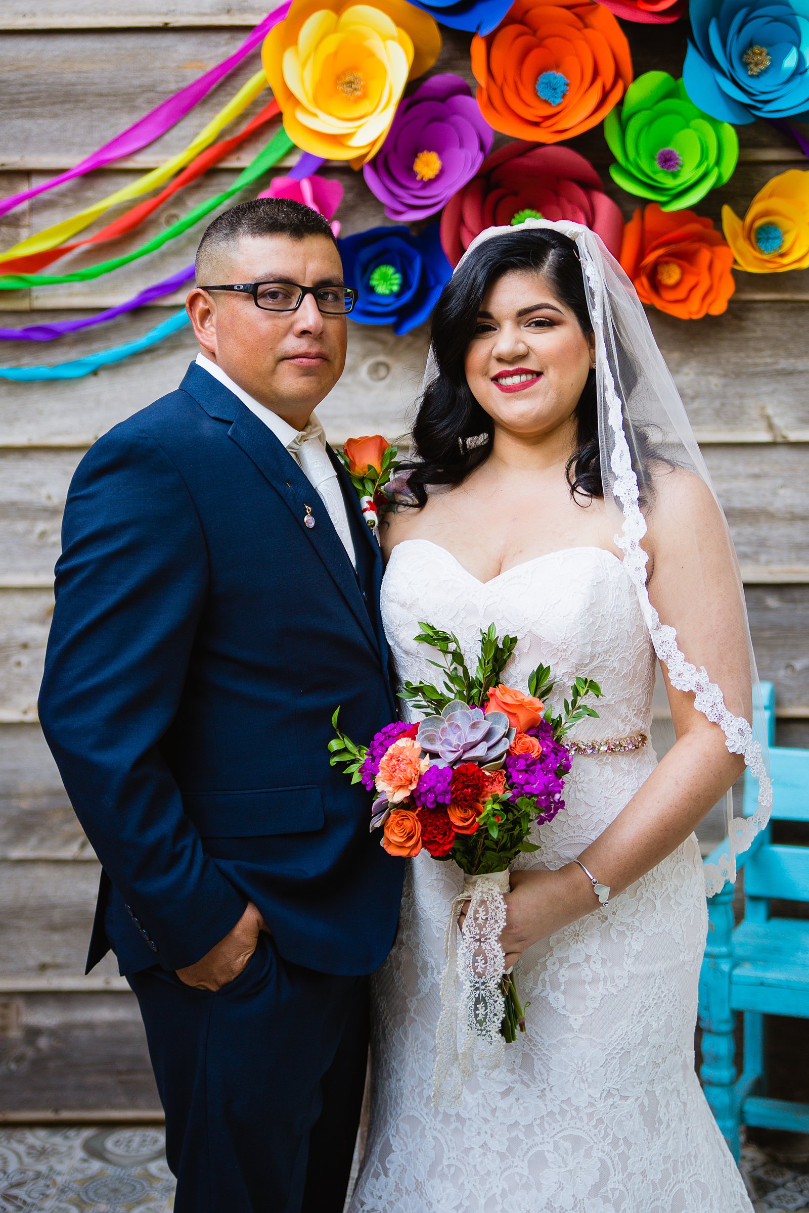 Bride and groom pose in front of a fiesta photo booth backdrop during their The Farmhouse at Schnepf Farms wedding by Arizona wedding photographer PMA Photography.