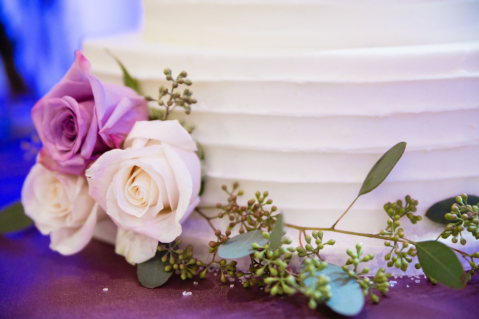 White and purple roses with eucalyptus decorating a simple white wedding cake by PMA Photography.