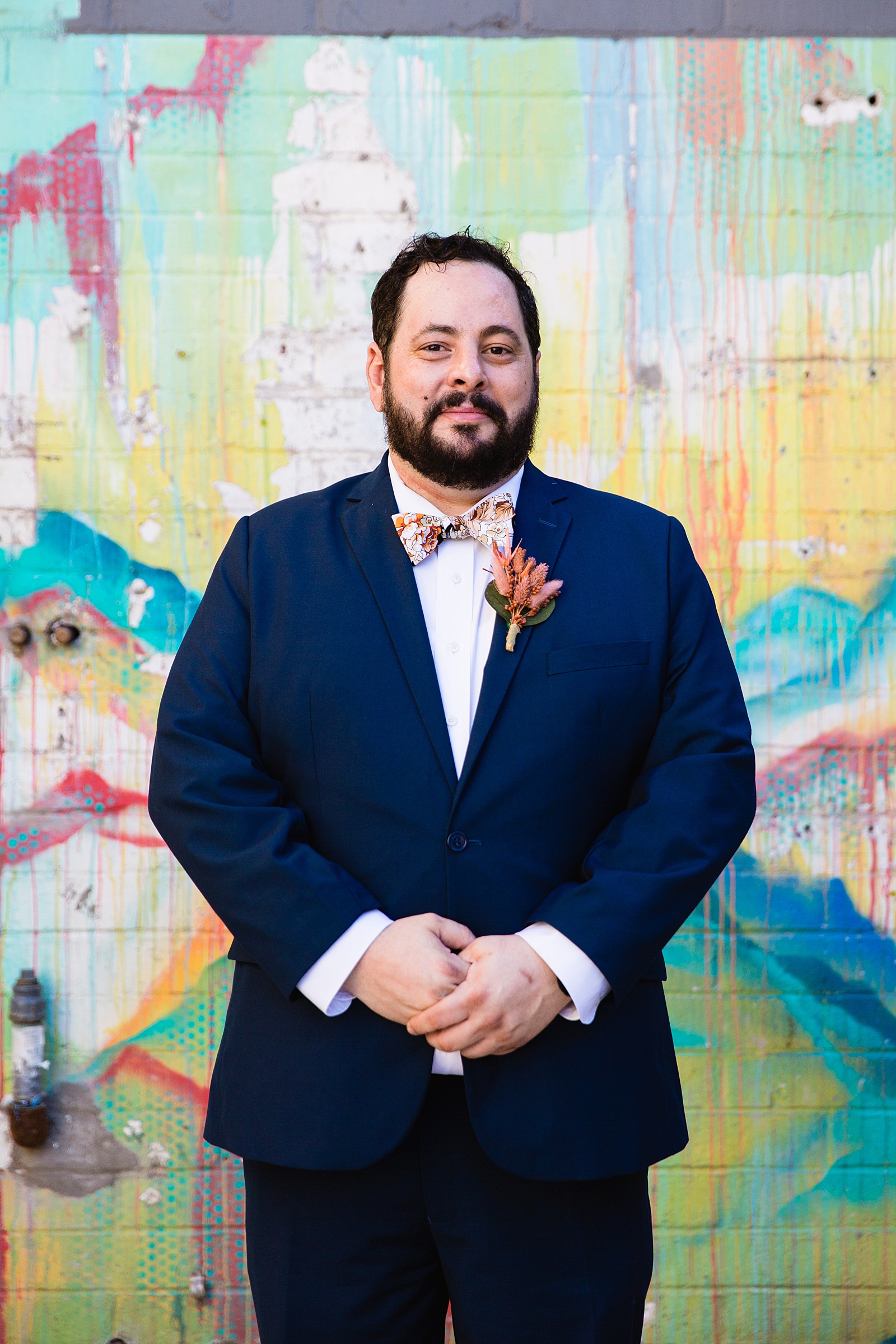 Groom in navy suit with floral bowtie in front of a colorful art mural for his MonOrchid wedding by PMA Photography.