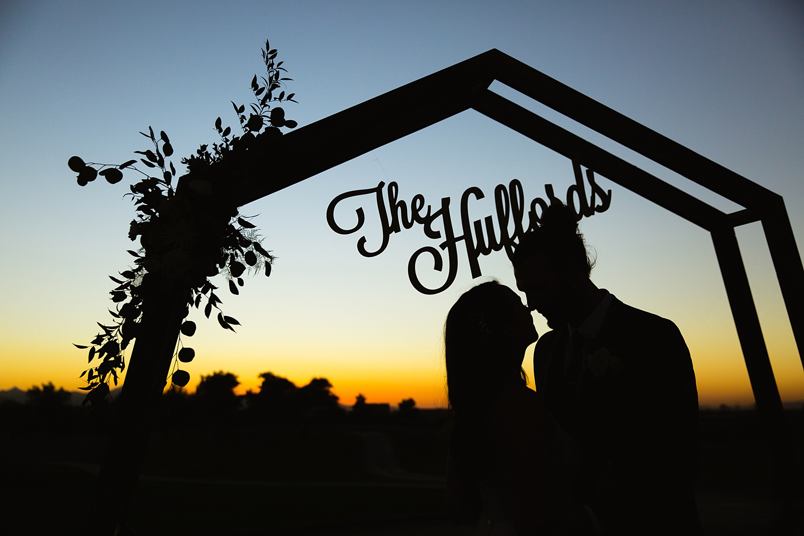 Silhouette of a bride and groom with their wedding arch and wood cut out of their last name with the sunset by Arizona wedding photographer PMA Photography.