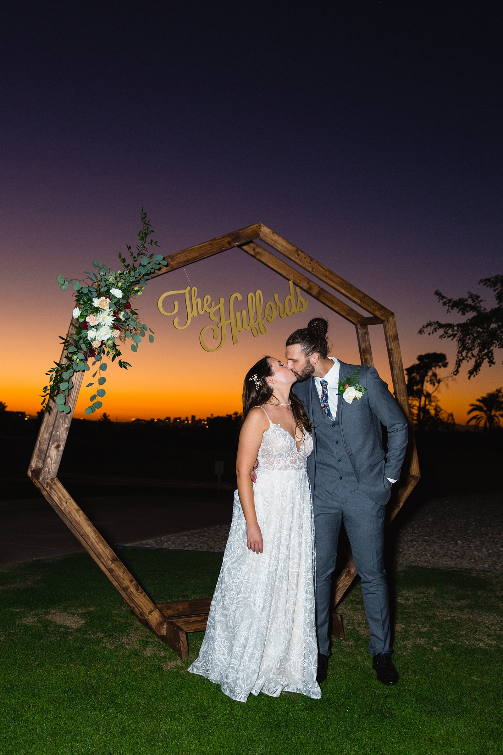 Couple share a kiss in front of their custom photo booth backdrop as the sun sets by Arizona wedding photographers PMA Photography.