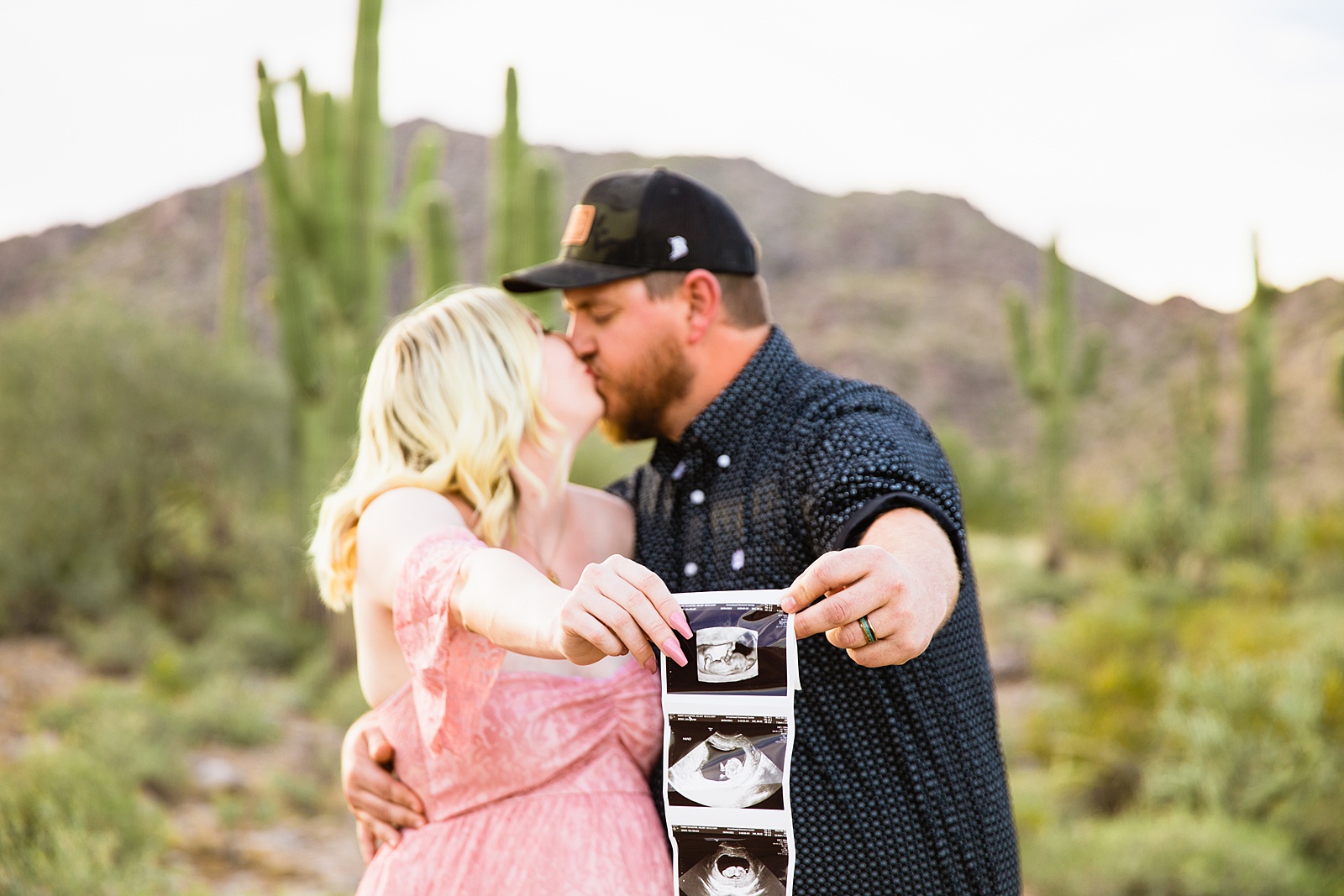 Couple kiss while holding out their ultrasound pictures by Arizona maternity photographer PMA Photography.